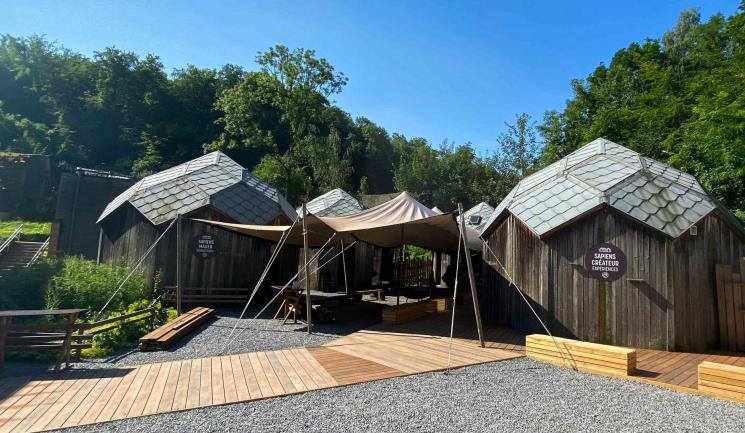 Cabanes d'exposition en bois avec rampe PMR en composite, toits en ardoise et voiles d'ombrage, au sein d'un espace aménagé avec du gravier et de la végétation